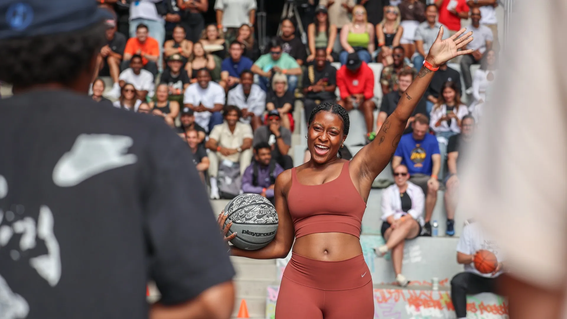 Một người phụ nữ mặc bộ đồ thể thao màu gỉ sắt, tay cầm quả bóng rổ và mỉm cười với đám đông đang ngồi.